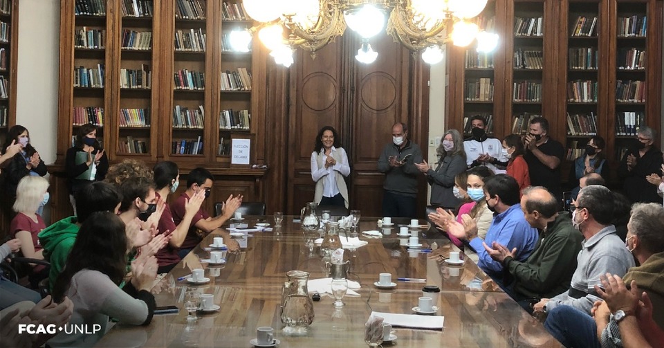 Esta imagen es una vista panorámica de la sesión extraordinaria de Consejo Directivo. Las y los Consejeros directivos están sentados alrededor de la mesa de la Biblioteca y el público alrededor. En la imagen se ve el ingreso de la Dra. Amalia Meza junto al Lic. Raúl Perdomo